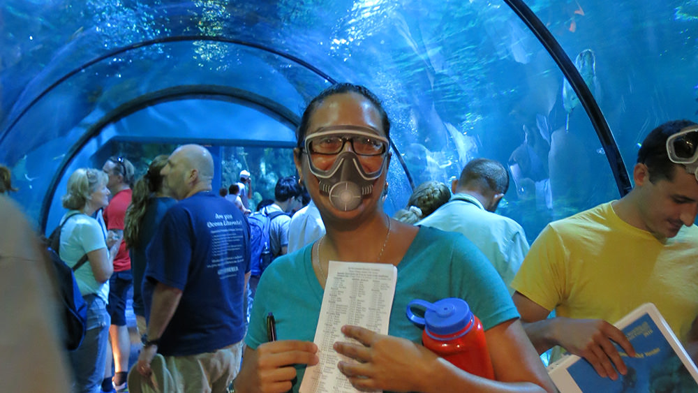 A woman wearing a paper scuba mask over her face while standing in a tunnel through an aquarium with a bunch of other adults. A man to her right has a paper scuba mask pushed up on his forehead. 