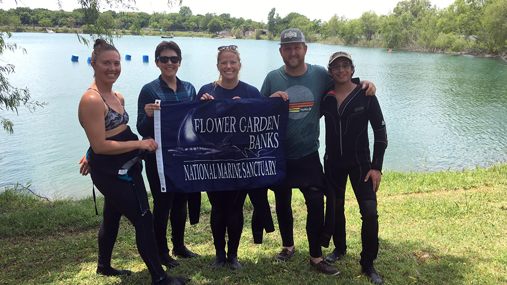 Five divers standing by a lake with a Flower Garden Banks National Marine Sanctuary banner