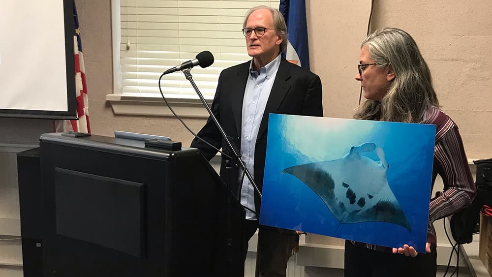 A man speaks at a podium as a woman holds a photo of a manta ray alongside.