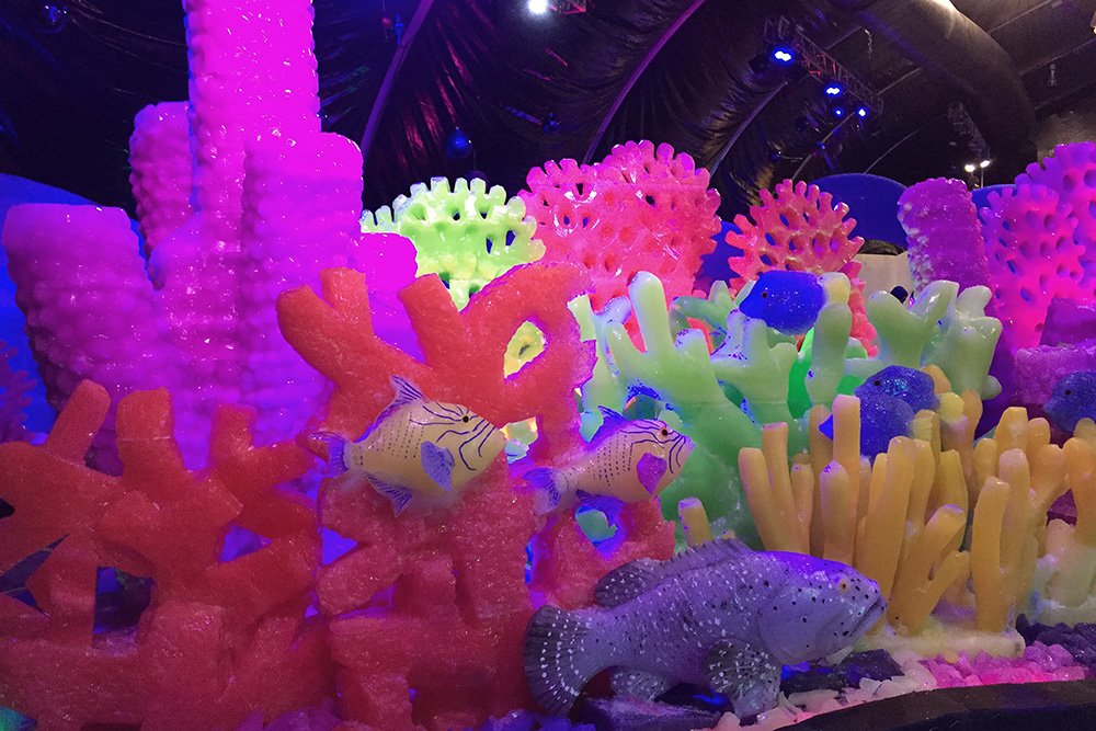 A colorful reef scene carved from ice. It includes bright red, pink, green and yellow sponges and coral, as well as colorful reef fish and a grouper.