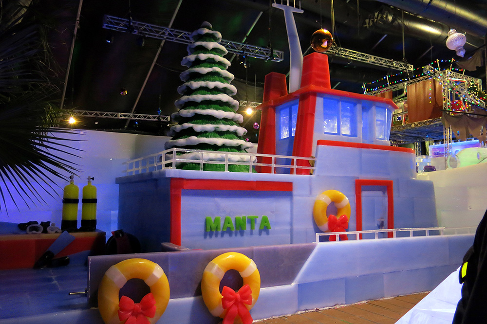 Replica of R/V MANTA carved from ice. the name MANTA is displayed on the side. Also displayed are life rings, and scuba gear all carved from ice. A large Christmas tree made from ice stands on the top deck.
