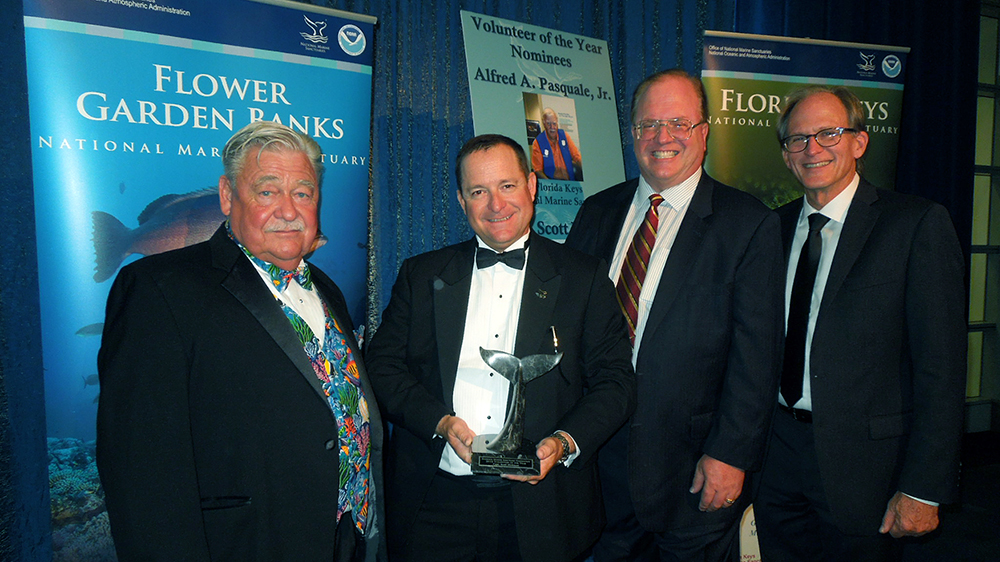 Left to right: Billy Causey, Scott Hickman holding a sculpted whale tail award, Clint Moore, G.P. Schmahl in front of the Flower Garden Banks NMS banner at an awards ceremony
