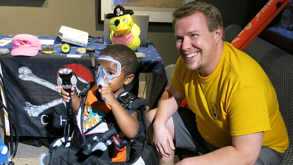 Adult volunteer squatting next to a child dressed in scuba gear and a paper dive mask