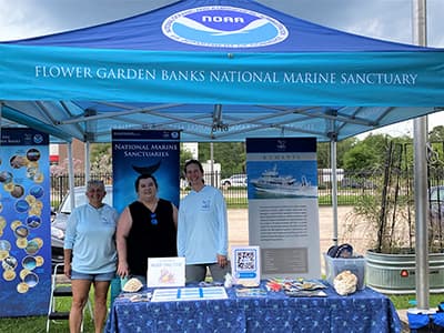 Three women stand in an outdoor booth with display banners and tabletop items about the sanctuary